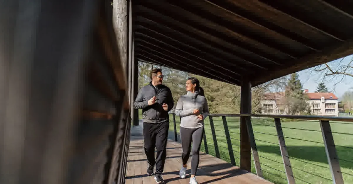 Middle-aged couple walking energetically through a sunlit park, revitalized and healthy, with a wellness center in the distance